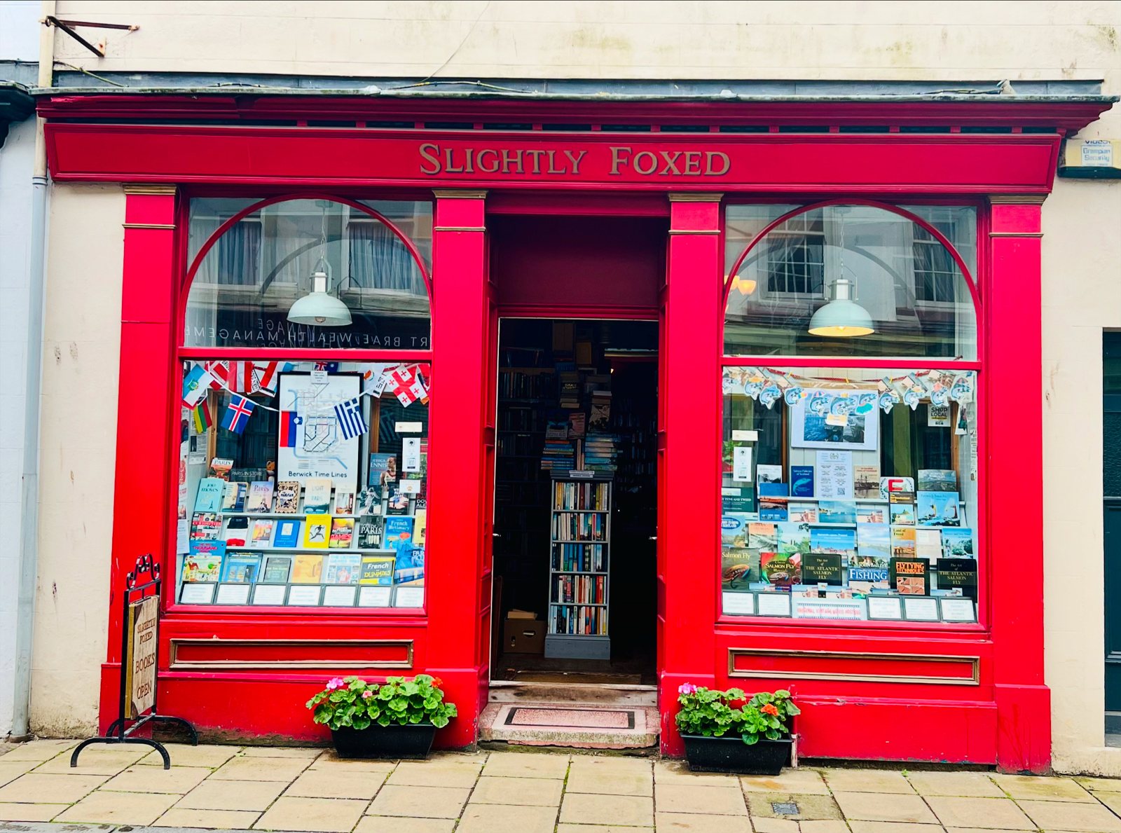 The red shopfront of Slightly Foxed at 31 Bridge Street, Berwick-upon-Tweed, with gilt lettering, arched windows full of books and geraniums on the pavement